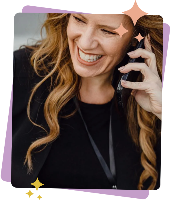 A woman with long, wavy hair is smiling while talking on the phone. She is wearing a black top and a black lanyard.